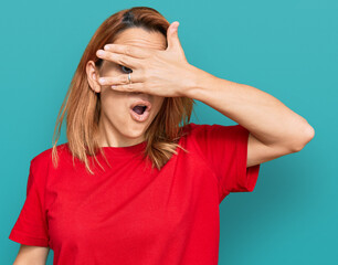 Hispanic young woman wearing casual red t shirt peeking in shock covering face and eyes with hand, looking through fingers with embarrassed expression.