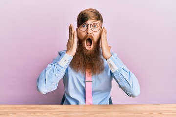 Young irish redhead man wearing business shirt and tie sitting on the table afraid and shocked,...