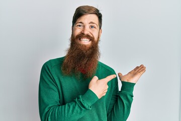 Redhead man with long beard wearing casual clothes amazed and smiling to the camera while presenting with hand and pointing with finger.