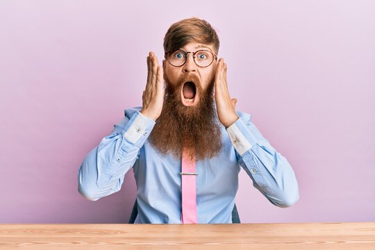 Young irish redhead man wearing business shirt and tie sitting on the table afraid and shocked, surprise and amazed expression with hands on face