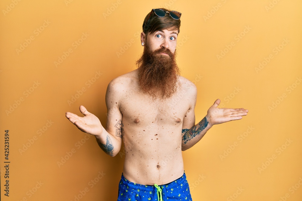 Wall mural redhead man with long beard wearing swimsuit and sunglasses clueless and confused expression with ar