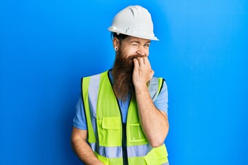 Redhead man with long beard wearing safety helmet and reflective jacket looking stressed and nervous with hands on mouth biting nails. anxiety problem.