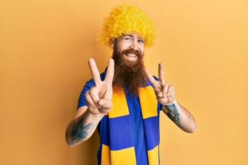 Redhead man with long beard football hooligan cheering game wearing funny wig smiling looking to the camera showing fingers doing victory sign. number two.