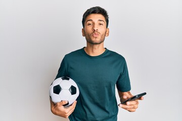 Young handsome man holding football ball looking at smartphone bet app looking at the camera blowing a kiss being lovely and sexy. love expression.