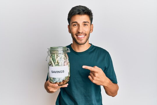 Young handsome man holding jar with savings smiling happy pointing with hand and finger