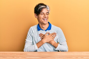 Fototapeta premium Young hispanic man wearing casual clothes sitting on the table smiling with hands on chest, eyes closed with grateful gesture on face. health concept.