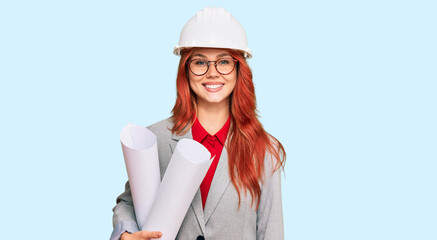 Young redhead architect woman wearing safety helmet holding blueprints looking positive and happy standing and smiling with a confident smile showing teeth