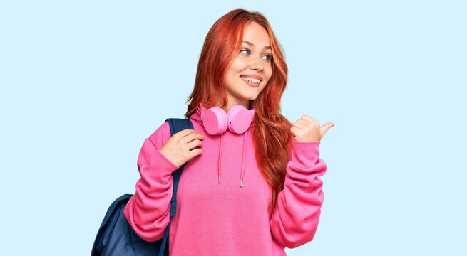 Young redhead woman holding student backpack and books pointing thumb up to the side smiling happy with open mouth