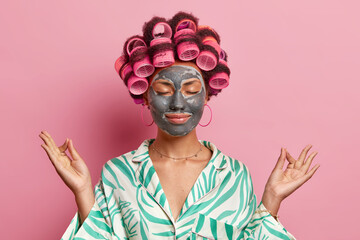 Relaxed female model meditates indoor makes zen gesture keeps eyes closed dressed in pajama applies beauty mask on face enjoys peacful domestic atmosphere isolated over pink background. Facial care
