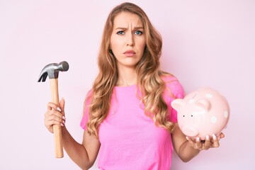 Young caucasian woman with blond hair holding piggy bank and hammer skeptic and nervous, frowning...