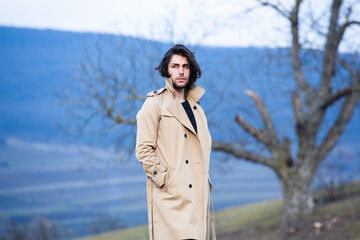 Image of young beautiful man with long hair wearing rain jacket in cold weather. Thinker european man in spring nature.