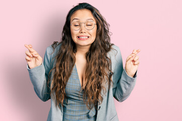 Young hispanic girl wearing business clothes and glasses gesturing finger crossed smiling with hope and eyes closed. luck and superstitious concept.