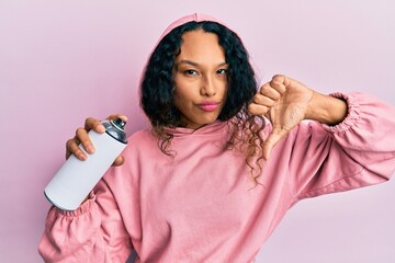 Young latin woman wearing sweatshirt holding graffiti spray with angry face, negative sign showing dislike with thumbs down, rejection concept
