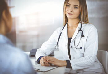 Young intelligent woman-doctor is listening to her patient's allergy's symptoms, while they are sitting together in the sunny cabinet in a clinic. Female physician, with a stethoscope, is writing at