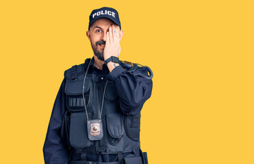 Young handsome man wearing police uniform covering one eye with hand, confident smile on face and surprise emotion.