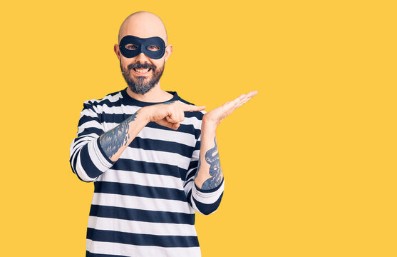 Young handsome man wearing burglar mask amazed and smiling to the camera while presenting with hand and pointing with finger.