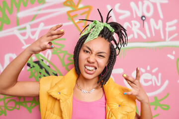 Adolescence street art and youth culture concept. Cheeky trendy hipster girl with dreadlocks makes cool gesture clenches golden teeth wears fashionable clothes poses against colorful graffiti wall. - obrazy, fototapety, plakaty