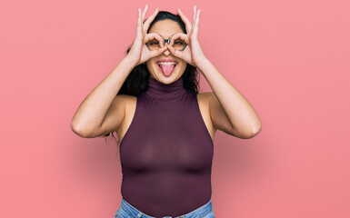 Brunette young woman wearing casual clothes and glasses doing ok gesture like binoculars sticking tongue out, eyes looking through fingers. crazy expression.