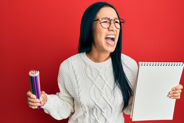 Beautiful hispanic woman holding canvas book and colored pencils angry and mad screaming frustrated and furious, shouting with anger looking up.