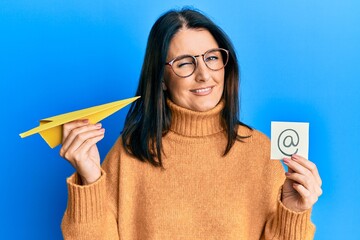 Middle age brunette woman holding email symbol and paper plane winking looking at the camera with...