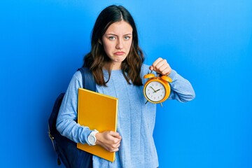 Young brunette student woman holding alarm clock skeptic and nervous, frowning upset because of...