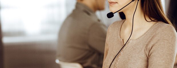 Young friendly girl in headsets is talking to a firm's client, while sitting at the desk in a modern office together with her colleague. Call center operators at work - obrazy, fototapety, plakaty