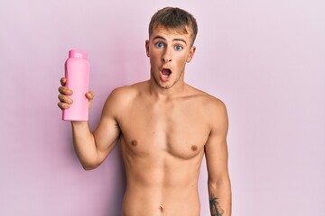 Fototapeta premium Young caucasian man shirtless holding shampoo bottle scared and amazed with open mouth for surprise, disbelief face