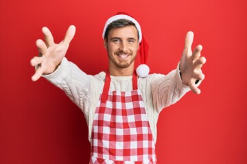 Handsome caucasian man wearing baker apron and christmas hat looking at the camera smiling with open arms for hug. cheerful expression embracing happiness.