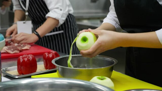 Two unrecognizable chefs are working in restaurant kitchen. One of them is peeling fresh green apple while another is cutting meat