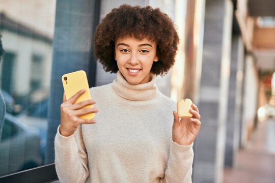 Young hispanic girl smiling happy using smartphone and holding earphones at the city.