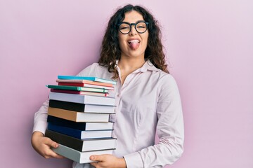 Young brunette woman with curly hair holding a pile of books sticking tongue out happy with funny expression.