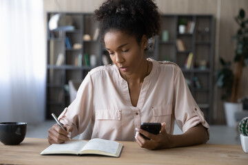 Focused young African American woman make notes handwrite in notebook use application on smartphone. Millennial biracial female write make list plan with cellphone. Planner, management concept. - obrazy, fototapety, plakaty