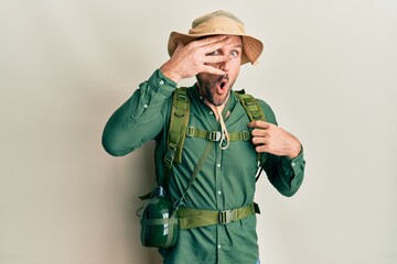 Handsome man with beard wearing explorer hat and backpack peeking in shock covering face and eyes with hand, looking through fingers with embarrassed expression.