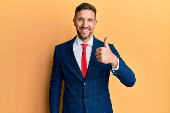 Handsome man with beard wearing business suit and tie smiling happy and positive, thumb up doing excellent and approval sign