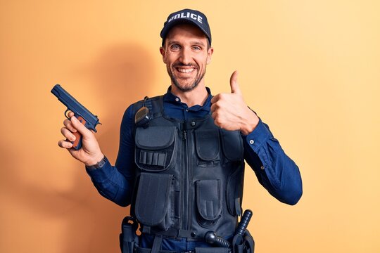 Handsome policeman wearing uniform and bulletprof holding gun over yellow background smiling happy and positive, thumb up doing excellent and approval sign