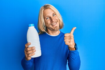 Caucasian young man with long hair holding liter bottle of milk smiling happy and positive, thumb up doing excellent and approval sign