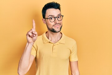 Fototapeta premium Young hispanic man wearing casual clothes and glasses showing and pointing up with finger number one while smiling confident and happy.