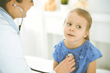 Obraz premium Doctor examining a child by stethoscope. Happy smiling girl patient dressed in blue dress is at usual medical inspection. Medicine concept