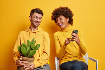 Happy young African American woman shows something funny on smartphone to boyfriend spend free time together sit on chairs isolated over yellow background. People technology lifestyle concept