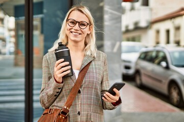Fototapeta premium Young blonde businesswoman using smartphone drinking coffee at the city.