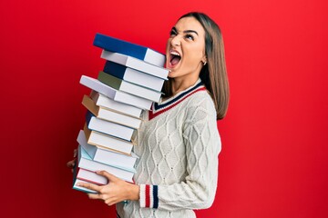 Young brunette student girl holding a pile of books angry and mad screaming frustrated and furious, shouting with anger looking up.