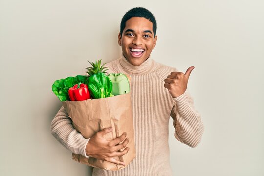 Young handsome hispanic man holding paper bag with groceries pointing thumb up to the side smiling happy with open mouth