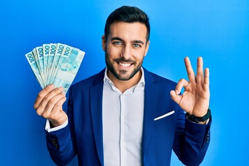 Young hispanic businessman wearing business suit holding brazilian real banknotes doing ok sign...