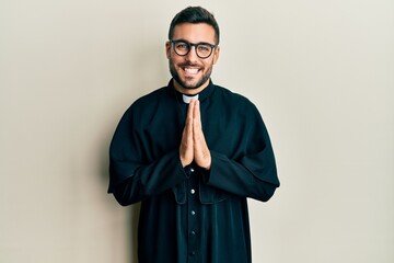 Obraz premium Young hispanic man wearing priest uniform standing over white background praying with hands together asking for forgiveness smiling confident.