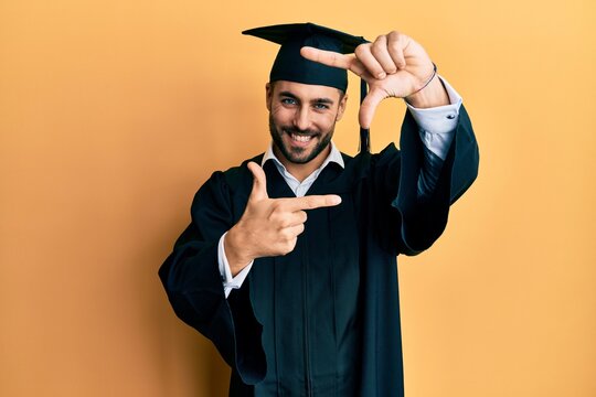 Young hispanic man wearing graduation cap and ceremony robe smiling making frame with hands and fingers with happy face. creativity and photography concept.