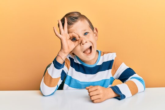 Adorable caucasian kid wearing casual clothes sitting on the table doing ok gesture with hand smiling, eye looking through fingers with happy face.