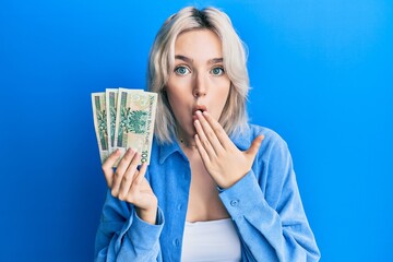 Young blonde girl holding 50 polish zloty banknotes covering mouth with hand, shocked and afraid for mistake. surprised expression