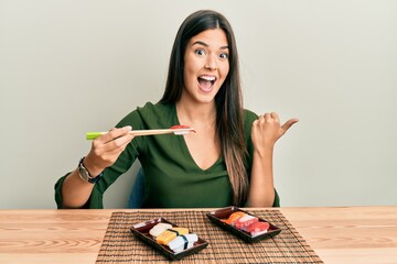 Young brunette woman eating sushi sitting on the table pointing thumb up to the side smiling happy...