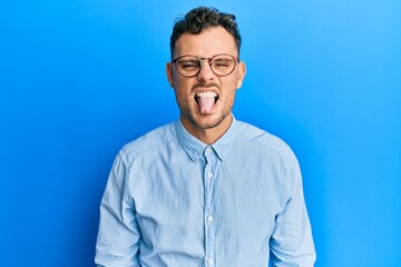 Young hispanic man wearing casual clothes and glasses sticking tongue out happy with funny expression. emotion concept.