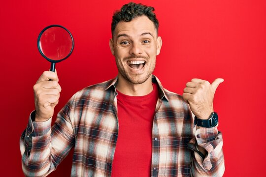 Young hispanic man holding magnifying glass looking for stain at clothes pointing thumb up to the side smiling happy with open mouth